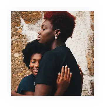 Two African American women dressed in black hugging in front of a stone wall