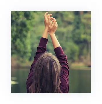 Woman at lake with purple top facing away from the camera with hands in the air in meditative pose